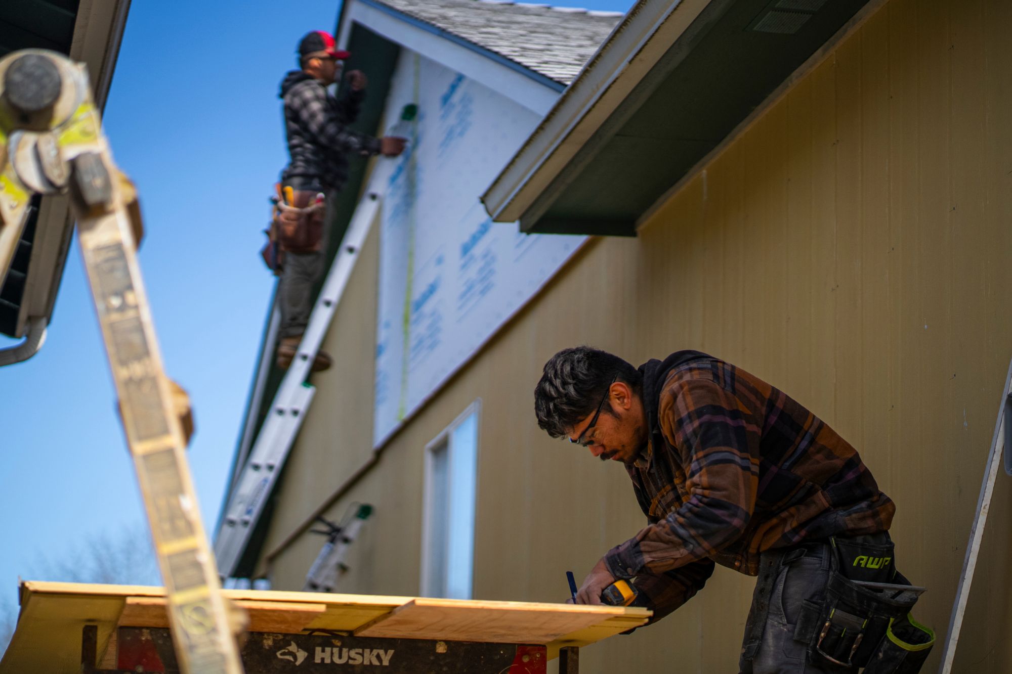 man in the foreground measuring boards while another assesses the siding in the background